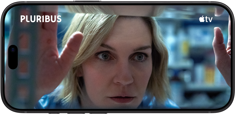 Un iPhone 18 Pro en orientation paysage, affichant un gros plan d’une femme qui regarde par une fenêtre. Titre de la série : Pluribus.