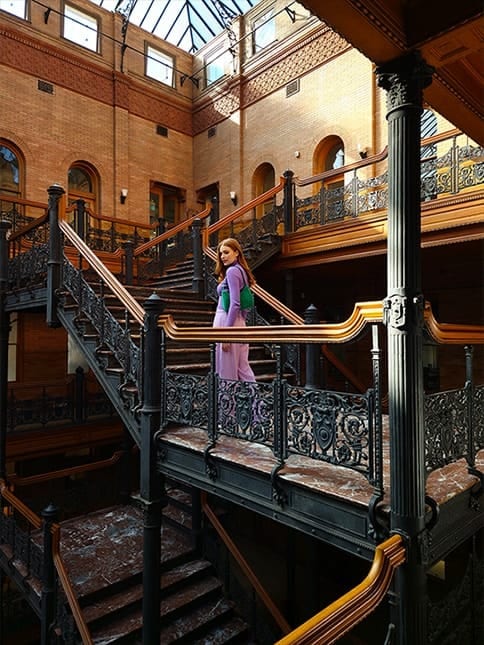 A high resolution photo of a woman standing in a brick atrium with multiple floors of stairs with intricately designed ironwork. Next, the same stairs from another angle and at a distance showing the architecture of the atrium. The 50 megapixel camera is able to capture the scene's rich detail and colour. Both photos were taken with Galaxy S23 plus using ISO 25, wide mode, and an aperture of F 1.8.