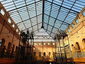 A high resolution photo of a woman standing in a brick atrium with multiple floors of stairs with intricately designed ironwork. Next, the same stairs from another angle and at a distance showing the architecture of the atrium. The 50 megapixel camera is able to capture the scene's rich detail and colour. Both photos were taken with Galaxy S23 plus using ISO 25, wide mode, and an aperture of F 1.8.