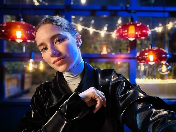 A woman takes a colourful, clear selfie in a dark setting with string lights and lanterns behind her. The photo was taken with Galaxy S23 Ultra using ISO 1600, wide mode, and an aperture of F 2.2