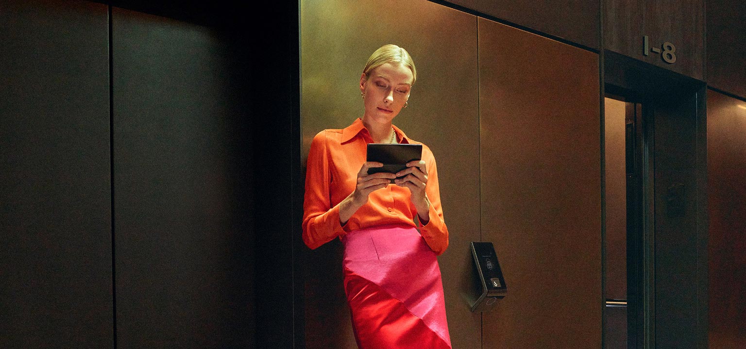 A woman stands by an elevator, holding an unfolded Galaxy Z Fold4 and looking at its Main Screen intently.
