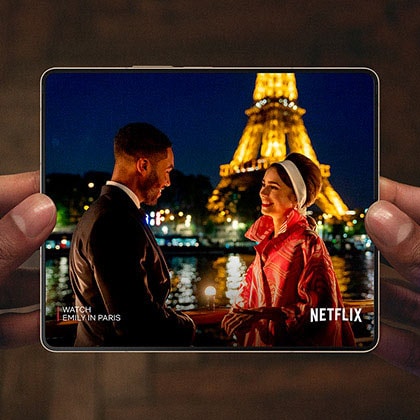 Hands holding an unfolded Galaxy Z Fold4. The Main Screen displays a scene from a Netflix video of a man and
						woman talking in front of the Eiffel Tower.
